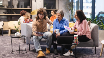 Indywidualne wsparcie, cyfrowe innowacje i inkluzyjne podejście – nowoczesny pracownik Publicznych Służb Zatrudnienia | Studia podyplomowe we Wrocławiu | Uniwersytet Dolnośląski DSW we Wrocławiu: Na zdjęciu trzy osoby siedzą na szarej sofie w nowoczesnym, jasnym wnętrzu. Młody mężczyzna w okularach, beżowej koszuli i dżinsach pokazuje coś na tablecie dwóm kobietom. Kobieta w niebieskiej koszuli trzyma kubek, a druga, w różowej bluzce, przegląda dokumenty. Przed nimi stoi stolik z laptopem i kubkiem. W tle widać więcej osób rozmawiających oraz dużą witrynę z dekoracjami. Atmosfera sprzyja pracy zespołowej i wymianie pomysłów.