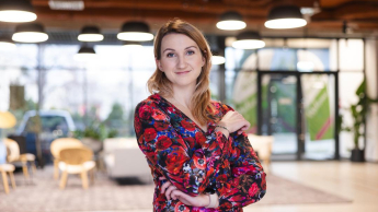 Psychologia skutecznej sprzedaży | Studia podyplomowe we Wrocławiu | Uniwersytet Dolnośląski DSW we Wrocławiu: Na zdjęciu kobieta w kolorowej, kwiecistej sukience stoi w nowoczesnym, jasnym wnętrzu. Uśmiecha się delikatnie i ma założone ręce. W tle widoczna jest przestrzeń z wygodnymi fotelami, dużymi oknami i dekoracyjnymi lampami sufitowymi. Atmosfera miejsca jest przyjazna i profesjonalna.