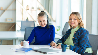 Automatyzacja procesów biznesowych | Studia podyplomowe we Wrocławiu | Uniwersytet Dolnośląski DSW we Wrocławiu: Na zdjęciu dwie młode kobiety siedzą przy stole w nowoczesnym, jasnym biurze. Jedna z nich uśmiecha się szeroko, trzymając kubek, druga patrzy spokojnie w stronę aparatu. Na stole leżą laptop, notatnik, dokumenty i kubki. W tle widać regał z dekoracjami i dużą ilość naturalnego światła wpadającego przez okna. Atmosfera jest przyjazna i profesjonalna.