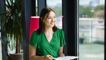 Pedagogika resocjalizacyjna z profilaktyką społeczną | Studia I stopnia we Wrocławiu | Uniwersytet Dolnośląski DSW we Wrocławiu: Kobieta w zielonej sukience stoi przy oknie z notesem w ręku. Uśmiecha się, spoglądając na zewnątrz. W tle widoczne są rośliny doniczkowe i lampka.