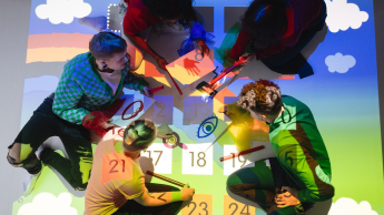 Pedagogika | Studia I stopnia we Wrocławiu | Uniwersytet Dolnośląski DSW we Wrocławiu: Pięć osób siedzi na podłodze wokół kolorowej interaktywnej projekcji, która przedstawia liczby, symbole i rysunki. Uczestnicy trzymają kolorowe pałeczki i kartki z ilustracjami dłoni, oczu i innych elementów. Scena sugeruje kreatywną pracę grupową lub zabawę edukacyjną.