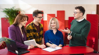 Zarządzanie | Studia I stopnia we Wrocławiu | Uniwersytet Dolnośląski DSW we Wrocławiu: Grupa studentów stoi przy czerwonym stole i rozmawia z wykładowcą. Jedna osoba robi notatki, inna trzyma książkę, a na stole leży otwarty laptop. W tle jasne wnętrze i czerwone panele akustyczne.