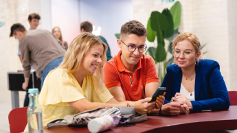 Marketing i social media | Studia I stopnia we Wrocławiu | Uniwersytet Dolnośląski DSW we Wrocławiu: „Trzy uśmiechnięte osoby siedzą przy stole i wspólnie przeglądają treści na telefonie w przestrzeni coworkingowej.”