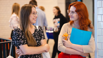 Zarządzanie | Studia I stopnia we Wrocławiu | Uniwersytet Dolnośląski DSW we Wrocławiu: Dwie studentki stoją przy ścianie, uśmiechają się i rozmawiają. Jedna trzyma segregator i butelkę wody, druga ma torbę na ramieniu.