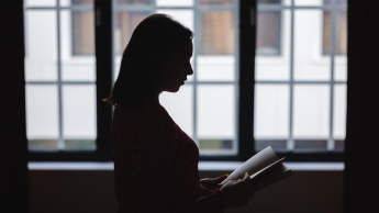 Psychologia kliniczna z psychoterapią | Studia jednolite magisterskie we Wrocławiu | Uniwersytet Dolnośląski DSW we Wrocławiu: Sylwetka kobiety z książką na tle dużych okien. Wnętrze jest zacienione, postać widoczna w półmroku.