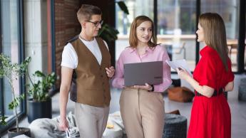 HR Business Partner z elementami coachingu | Studia I stopnia we Wrocławiu | Uniwersytet Dolnośląski DSW we Wrocławiu: Na zdjęciu widzimy trzy osoby rozmawiające w nowoczesnym biurze. Kobieta po lewej trzyma laptopa, ubrana jest w różową koszulę i beżowe spodnie. Mężczyzna obok niej nosi okulary, biały T-shirt i brązową kamizelkę, trzyma kartkę papieru. Kobieta po prawej stronie ubrana jest w czerwoną sukienkę i trzyma dokumenty. W tle widać duże okna, rośliny w doniczkach i wygodne siedziska, co tworzy przyjazną atmosferę pracy zespołowej.