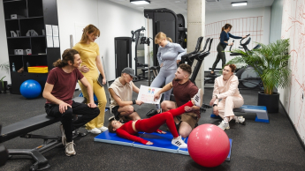 Sport | Studia I stopnia we Wrocławiu | Uniwersytet Dolnośląski DSW we Wrocławiu: zajęcia sportowe na siłowni, studenci oglądają wykonywanie ćwiczeń przez studentkę