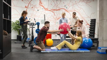 Trener przygotowania motorycznego | Studia I stopnia we Wrocławiu | Uniwersytet Dolnośląski DSW we Wrocławiu: zajęcia sportowe, studenci ćwiczą na macie podnoszenie piłek i skłony