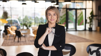 Psychologia kryzysu i interwencji kryzysowej | Uniwersytet Dolnośląski DSW we Wrocławiu: Kobieta w eleganckim stroju stoi w nowoczesnym wnętrzu, trzymając okulary i patrząc z uśmiechem w obiektyw.