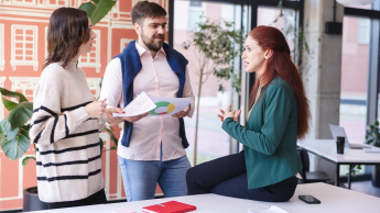 Psychologia zarządzania i biznesu | Studia jednolite magisterskie we Wrocławiu | Uniwersytet Dolnośląski DSW we Wrocławiu: Trzy osoby rozmawiają w jasnym, nowoczesnym biurze – kobieta siedzi na biurku, pozostali stoją z dokumentami w dłoniach.