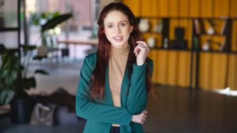 Psychologia dzieci i młodzieży | Studia jednolite magisterskie we Wrocławiu | Uniwersytet Dolnośląski DSW we Wrocławiu: Młoda kobieta z rudymi włosami uśmiecha się do obiektywu w przestrzeni coworkingowej.