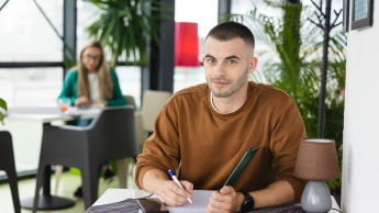 Mężczyzna w brązowej bluzie pisze coś w zeszycie, siedząc przy stoliku w kawiarni z roślinami.