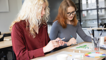 Wyniki wyszukiwania | Uniwersytet Dolnośląski DSW we Wrocławiu: Dwie studentki malujące przy stoliku w pracowni – jedna w bordowej koszuli, druga w szarej bluzce.