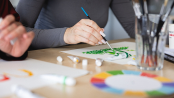 Edukacja i rehabilitacja osób z niepełnosprawnością intelektualną – dla pedagogów specjalnych | Studia podyplomowe we Wrocławiu | Uniwersytet Dolnośląski DSW we Wrocławiu: Zbliżenie na dłonie osoby malującej zielony wzór farbą na papierze, w otoczeniu przyborów plastycznych.