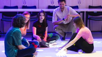Pedagogika opiekuńczo-wychowawcza z elementami wielokulturowości | Studia I stopnia we Wrocławiu | Uniwersytet Dolnośląski DSW we Wrocławiu: Grupa młodych dorosłych siedzi na podłodze w sali edukacyjnej, uczestnicząc w zajęciach z wykorzystaniem interaktywnej podłogi sensorycznej.