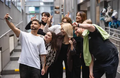 Aktualności | Uniwersytet Dolnośląski DSW we Wrocławiu: Grupa młodych osób pozuje do wspólnego selfie w jasnym korytarzu uczelni. Wszyscy uśmiechają się, robią gesty pokoju i radośnie spoglądają w stronę telefonu trzymanego przez mężczyznę z przodu.