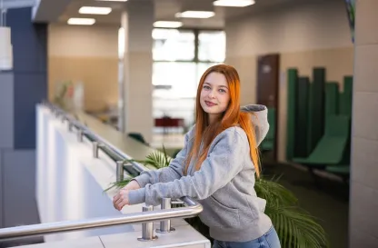 Aktualności | Uniwersytet Dolnośląski DSW we Wrocławiu: studentka w szarej bluzie i rudych włosach, opierająca się o barierkę