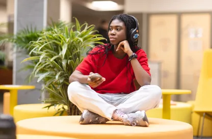 Aktualności | Uniwersytet Dolnośląski DSW we Wrocławiu: Studentka w czerwonej bluzie i słuchawkach siedzi po turecku na żółtym siedzisku, trzyma telefon.