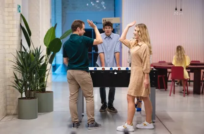 Aktualności | Uniwersytet Dolnośląski DSW we Wrocławiu: Dwoje studentów przybija sobie piątkę po grze w piłkarzyki, trzeci student patrzy na nich z uśmiechem.