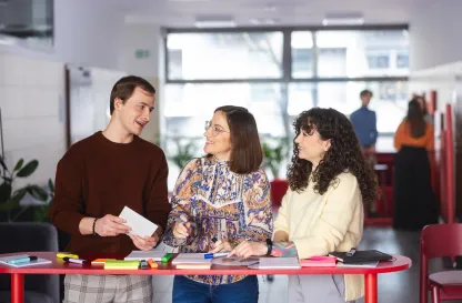 Poradniki - dzielimy się wiedzą | Uniwersytet Dolnośląski DSW we Wrocławiu: studenci stojący przy czerwonym stole z notatkami