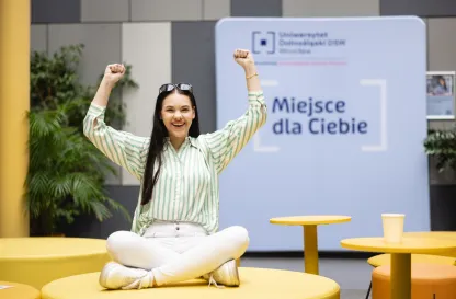Poradniki - dzielimy się wiedzą | Uniwersytet Dolnośląski DSW we Wrocławiu: Grafika główna: Uniwersytet Dolnośląski DSW, dla nowych studentów, studentka siedzi na sofie i trzyma ręce w górze w geście radości, w tyle napis na ścianie "Miejsce dla ciebie"