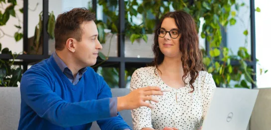 Studia II stopnia we Wrocławiu | Uniwersytet Dolnośląski DSW we Wrocławiu: Mężczyzna w niebieskim swetrze i kobieta w okularach pracują razem przy stole z laptopem w nowoczesnym wnętrzu z roślinami w tle. Oboje rozmawiają i analizują notatki.