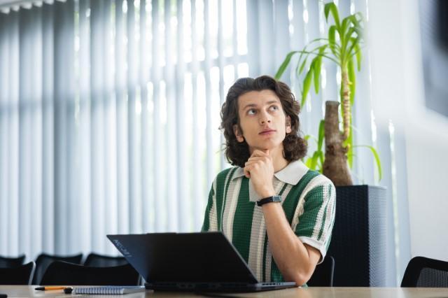 Druk 3D w przemyśle | Studia I stopnia we Wrocławiu | Uniwersytet Dolnośląski DSW we Wrocławiu: Młody mężczyzna z długimi, ciemnymi włosami siedzi przy stole z laptopem. Opiera brodę na dłoni, patrząc w górę z zamyśloną miną. W tle roślina doniczkowa i pionowe żaluzje w oknie.