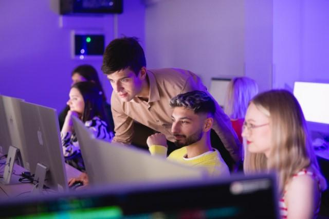 Motion design i animacja w marketingu | Studia I stopnia we Wrocławiu | Uniwersytet Dolnośląski DSW we Wrocławiu: Grupa studentów pracuje przy komputerach w sali komputerowej o fioletowym oświetleniu. Mężczyzna w beżowej koszuli pochyla się, pomagając koledze w żółtym swetrze. W tle dwie kobiety skupione na ekranach.
