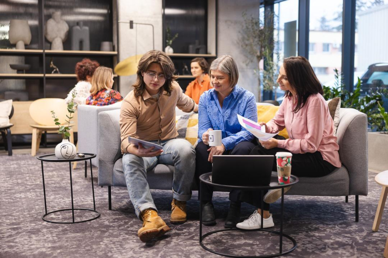 Growth Performance Marketing | Studia podyplomowe we Wrocławiu | Uniwersytet Dolnośląski DSW we Wrocławiu: Na zdjęciu trzy osoby siedzą na szarej sofie w nowoczesnym, jasnym wnętrzu. Młody mężczyzna w okularach, beżowej koszuli i dżinsach pokazuje coś na tablecie dwóm kobietom. Kobieta w niebieskiej koszuli trzyma kubek, a druga, w różowej bluzce, przegląda dokumenty. Przed nimi stoi stolik z laptopem i kubkiem. W tle widać więcej osób rozmawiających oraz dużą witrynę z dekoracjami. Atmosfera sprzyja pracy zespołowej i wymianie pomysłów.