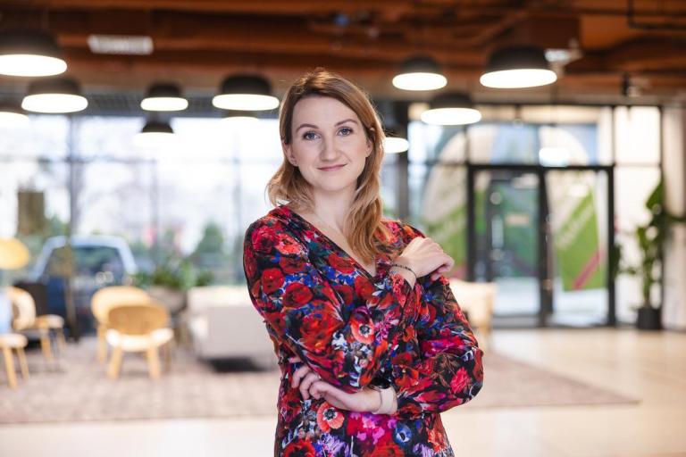 Dział Studiów Podyplomowych i Szkoleń | Uniwersytet Dolnośląski DSW we Wrocławiu: Na zdjęciu kobieta w kolorowej, kwiecistej sukience stoi w nowoczesnym, jasnym wnętrzu. Uśmiecha się delikatnie i ma założone ręce. W tle widoczna jest przestrzeń z wygodnymi fotelami, dużymi oknami i dekoracyjnymi lampami sufitowymi. Atmosfera miejsca jest przyjazna i profesjonalna.