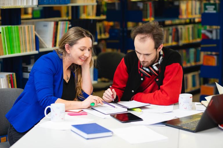 Biblioteka | Uniwersytet Dolnośląski DSW we Wrocławiu: Na zdjęciu dwoje studentów pracuje razem przy stole w bibliotece. Kobieta w niebieskiej marynarce uśmiecha się, patrząc na notatki, natomiast mężczyzna w czerwonej bluzie z czarnymi rękawami i słuchawkami na szyi zapisuje coś w zeszycie. Na stole znajdują się książki, zeszyty, laptop, tablet, kolorowe zakreślacze i kubki. W tle widać regały pełne książek. Atmosfera jest skupiona i sprzyja współpracy.