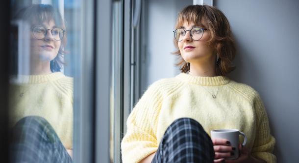 Interactive learning game for refugee and migrANT children's inclUsion and integRAtion (ANTURA) | Uniwersytet Dolnośląski DSW we Wrocławiu: Młoda kobieta w okularach i żółtym swetrze siedzi przy oknie z kubkiem w dłoni, patrząc zamyślona na zewnątrz. W oknie odbija się jej postać.