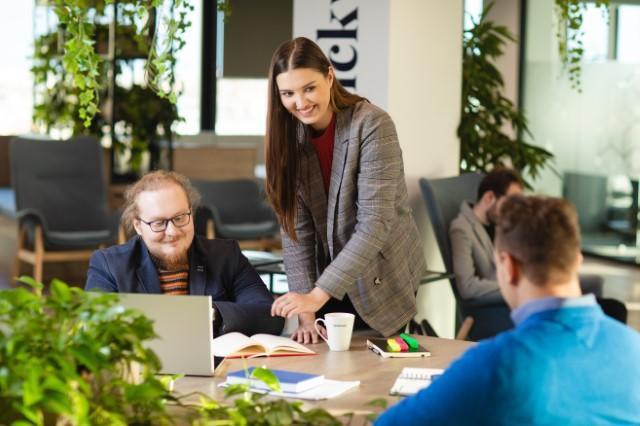 Studia podyplomowe we Wrocławiu | Uniwersytet Dolnośląski DSW we Wrocławiu: Trzy osoby rozmawiają i pracują przy wspólnym stole w nowoczesnym, zielonym biurze. Jedna kobieta stoi i uśmiecha się, wskazując na laptop, obok dwóch mężczyzn siedzących przy biurku.