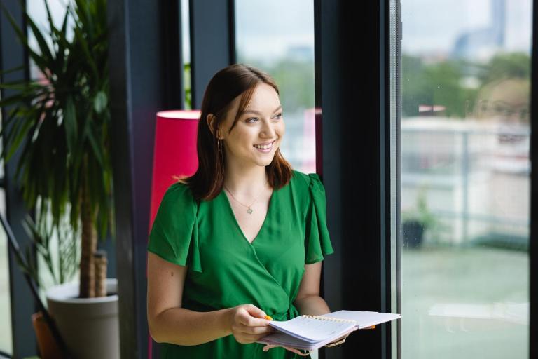 Pedagogika | Studia I stopnia we Wrocławiu | Uniwersytet Dolnośląski DSW we Wrocławiu: Kobieta w zielonej sukience stoi przy oknie z notesem w ręku. Uśmiecha się, spoglądając na zewnątrz. W tle widoczne są rośliny doniczkowe i lampka.