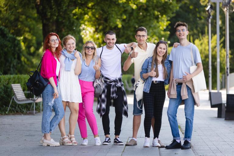 Pedagogika | Studia I stopnia we Wrocławiu | Uniwersytet Dolnośląski DSW we Wrocławiu: Grupa uśmiechniętych młodych ludzi pozuje razem na tle zieleni, stojąc blisko siebie i pokazując kciuki w górę. Panuje radosna, przyjazna atmosfera.