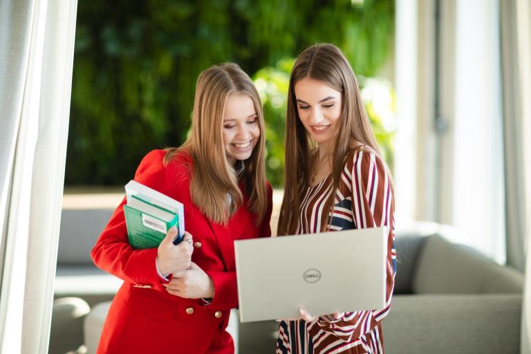 Pedagogika resocjalizacyjna z profilaktyką społeczną | Studia I stopnia we Wrocławiu | Uniwersytet Dolnośląski DSW we Wrocławiu: Dwie młode kobiety uśmiechają się, patrząc na ekran laptopa. Jedna z nich trzyma kilka książek. Ujęcie sugeruje wspólną naukę lub pracę nad projektem w przyjaznej atmosferze, w jasnym, zielonym otoczeniu.