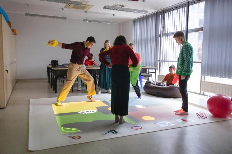 Pedagogika opiekuńczo-wychowawcza z elementami socjoterapii | Studia I stopnia we Wrocławiu | Uniwersytet Dolnośląski DSW we Wrocławiu: Kilka osób uczestniczy w ćwiczeniach na interaktywnej macie w sali dydaktycznej. Na podłodze wyświetlane są kolorowe symbole i pola, a uczestnicy poruszają się aktywnie, angażując się w zabawę edukacyjną.