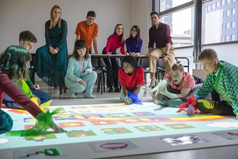 Terapia pedagogiczna z elementami arteterapii | Studia I stopnia we Wrocławiu | Uniwersytet Dolnośląski DSW we Wrocławiu: Grupa studentów i wykładowców bierze udział w zajęciach z wykorzystaniem maty interaktywnej. Część osób klęczy lub siedzi na podłodze, wskazując elementy wyświetlane na macie, inni obserwują z uśmiechami w tle. Atmosfera sprzyja zaangażowanej nauce i zabawie.