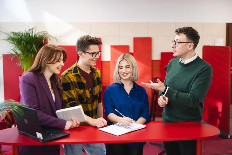 Zarządzanie zasobami ludzkimi | Studia I stopnia we Wrocławiu | Uniwersytet Dolnośląski DSW we Wrocławiu: Grupa studentów stoi przy czerwonym stole i rozmawia z wykładowcą. Jedna osoba robi notatki, inna trzyma książkę, a na stole leży otwarty laptop. W tle jasne wnętrze i czerwone panele akustyczne.