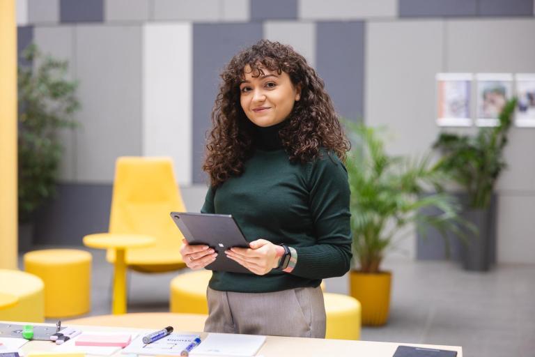 Zarządzanie | Studia I stopnia we Wrocławiu | Uniwersytet Dolnośląski DSW we Wrocławiu: Kobieta z kręconymi włosami w zielonym golfie trzyma tablet i uśmiecha się lekko. Za nią kolorowe, nowoczesne wnętrze z żółtymi meblami i roślinami. Na stole widać dokumenty i przybory do pisania.