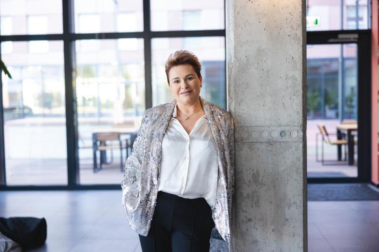 Zarządzanie zasobami ludzkimi | Studia I stopnia we Wrocławiu | Uniwersytet Dolnośląski DSW we Wrocławiu: „Kobieta w błyszczącej marynarce i białej koszuli opiera się o betonowy filar w nowoczesnym wnętrzu z dużymi oknami.”