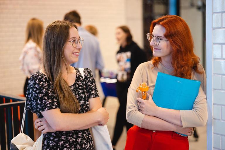 Leadership in business | Studia I stopnia we Wrocławiu | Uniwersytet Dolnośląski DSW we Wrocławiu: Dwie studentki stoją przy ścianie, uśmiechają się i rozmawiają. Jedna trzyma segregator i butelkę wody, druga ma torbę na ramieniu.