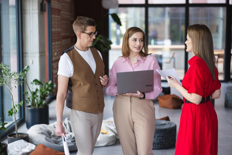 Strategie PR | Studia I stopnia we Wrocławiu | Uniwersytet Dolnośląski DSW we Wrocławiu: Troje pracowników lub studentów rozmawia w nowoczesnym wnętrzu biurowym. Jedna osoba trzyma laptopa, inna dokumenty.