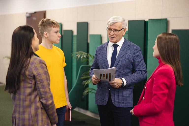 Zarządzanie i raportowanie w obszarze ESG | Studia I stopnia we Wrocławiu | Uniwersytet Dolnośląski DSW we Wrocławiu: Starszy wykładowca w garniturze rozmawia z trzema studentami. Trzyma w rękach dokumenty z kolorowymi wykresami. Wszyscy stoją w nowoczesnym wnętrzu z zielonymi dekoracjami ściennymi.
