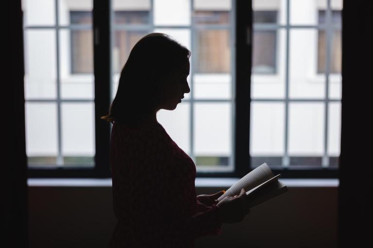 Psychologia w biznesie | Studia I stopnia we Wrocławiu | Uniwersytet Dolnośląski DSW we Wrocławiu: Sylwetka kobiety z książką na tle dużych okien. Wnętrze jest zacienione, postać widoczna w półmroku.