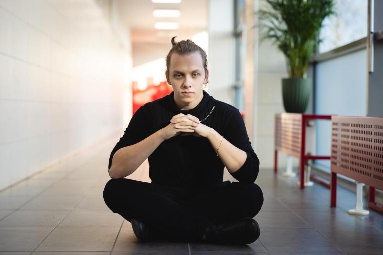 Psychologia w biznesie | Studia I stopnia we Wrocławiu | Uniwersytet Dolnośląski DSW we Wrocławiu: Młody mężczyzna w czarnym stroju siedzi po turecku na podłodze jasnego korytarza uczelni, patrząc prosto w obiektyw z złożonymi rękami, wyrażając pewność siebie i skupienie.