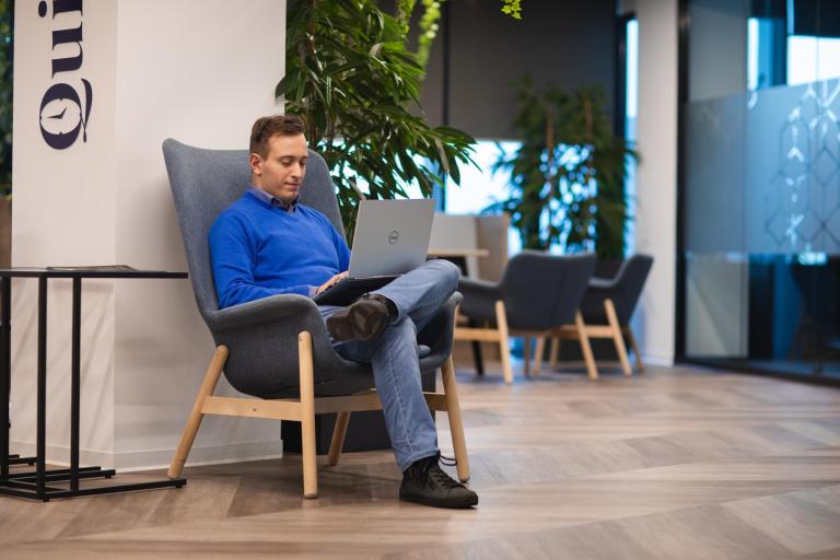 Psychologia marketingu i nowych mediów | Studia I stopnia we Wrocławiu | Uniwersytet Dolnośląski DSW we Wrocławiu: Mężczyzna w niebieskim swetrze i dżinsach siedzi wygodnie w fotelu z laptopem na kolanach. Pracuje w jasnej, nowoczesnej przestrzeni biurowej z dużą ilością roślin.