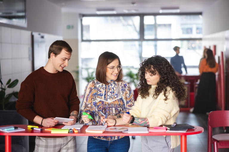 Human resources i coaching | Studia II stopnia we Wrocławiu | Uniwersytet Dolnośląski DSW we Wrocławiu: studenci stojący przy czerwonym stole, wykonują zadania projektowe