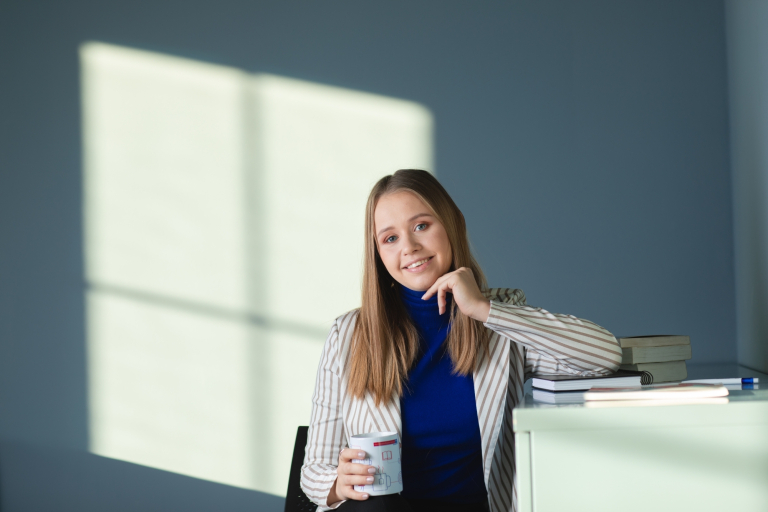Kreacja multimedialna i komunikacja wizualna | Studia I stopnia we Wrocławiu | Uniwersytet Dolnośląski DSW we Wrocławiu: uśmiechnięta studentka w niebieskim golfie i marynarce w paski, opiera się o biurko