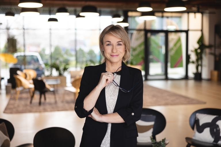 Psychologia kliniczna z psychoterapią | Studia jednolite magisterskie we Wrocławiu | Uniwersytet Dolnośląski DSW we Wrocławiu: Kobieta w eleganckim stroju stoi w nowoczesnym wnętrzu, trzymając okulary i patrząc z uśmiechem w obiektyw.